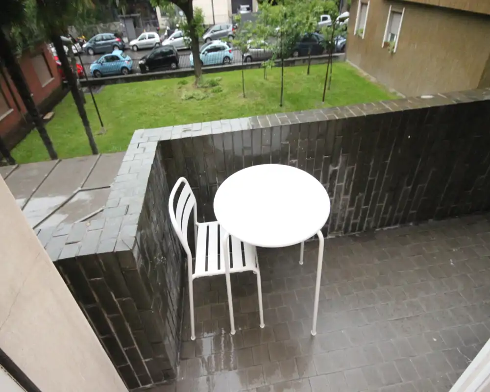 Piccolo balcone con tavolo rotondo bianco e una sedia abbinata che si affaccia su un cortile erboso; il pavimento piastrellato bagnato indica pioggia recente.