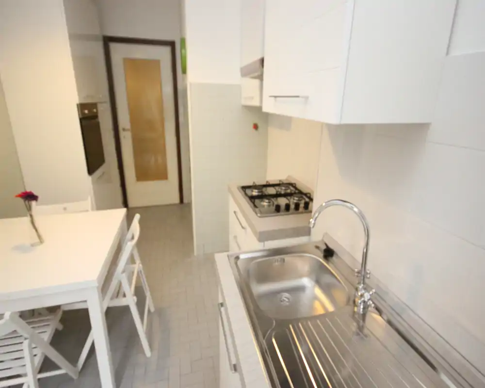 Cucina compatta e luminosa con lavello in acciaio, piano cottura a quattro fuochi e piccolo tavolo da pranzo. Pensili bianchi e pavimento piastrellato rendono lo spazio pulito e funzionale.