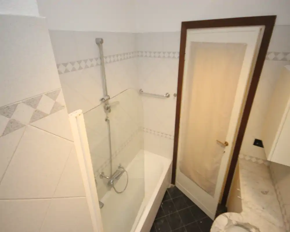 Bagno piastrellato e pulito con vasca, parete doccia in vetro, doccia a muro e porta accanto alla vasca. Piastrelle con bordo decorativo e pavimento scuro a contrasto con le pareti chiare.