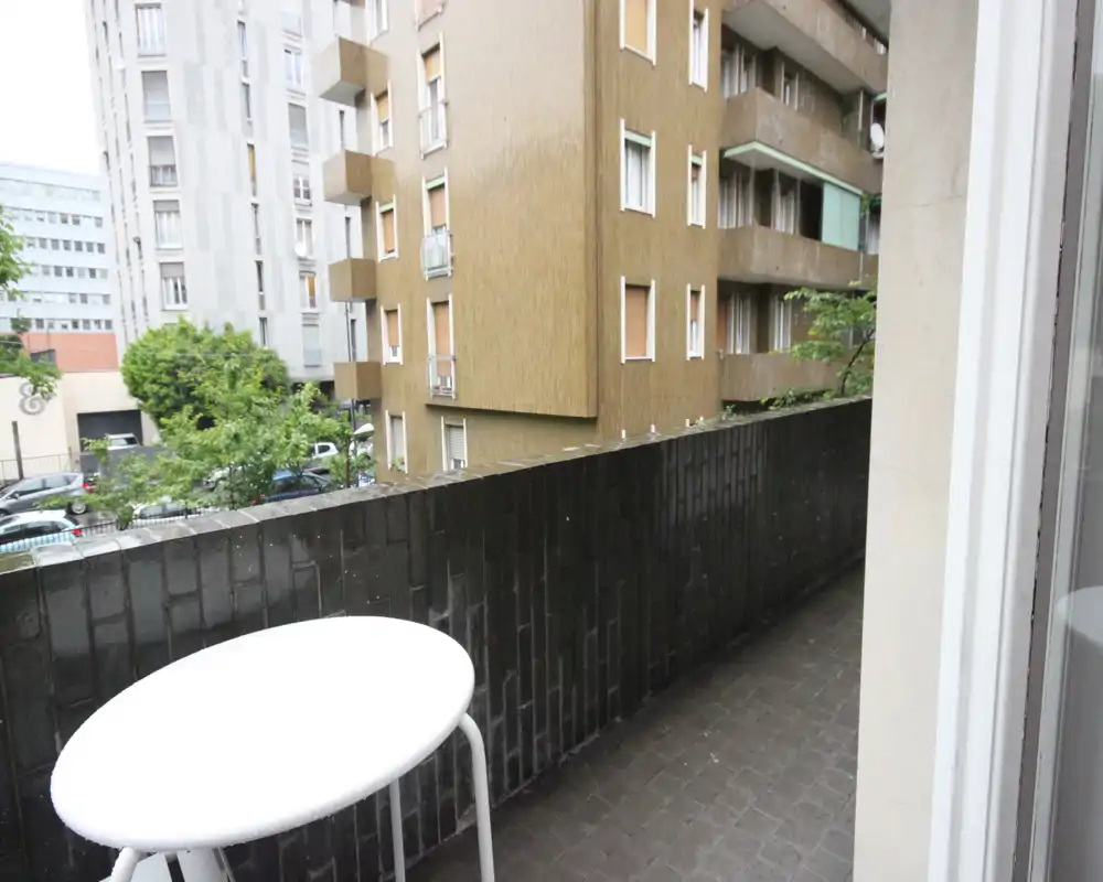 Balcone urbano con pavimento piastrellato e parapetto in mattoni bassi; è visibile un tavolino bianco rotondo e la vista si affaccia su edifici residenziali e piante a livello strada.