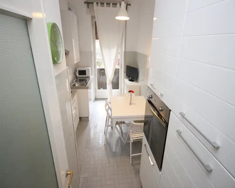 Cucina luminosa e stretta con elettrodomestici incassati, piccolo tavolo da pranzo e porta sul balcone. Pensili bianchi puliti, microonde e forno visibili; le tende chiare rendono l'ambiente accogliente.