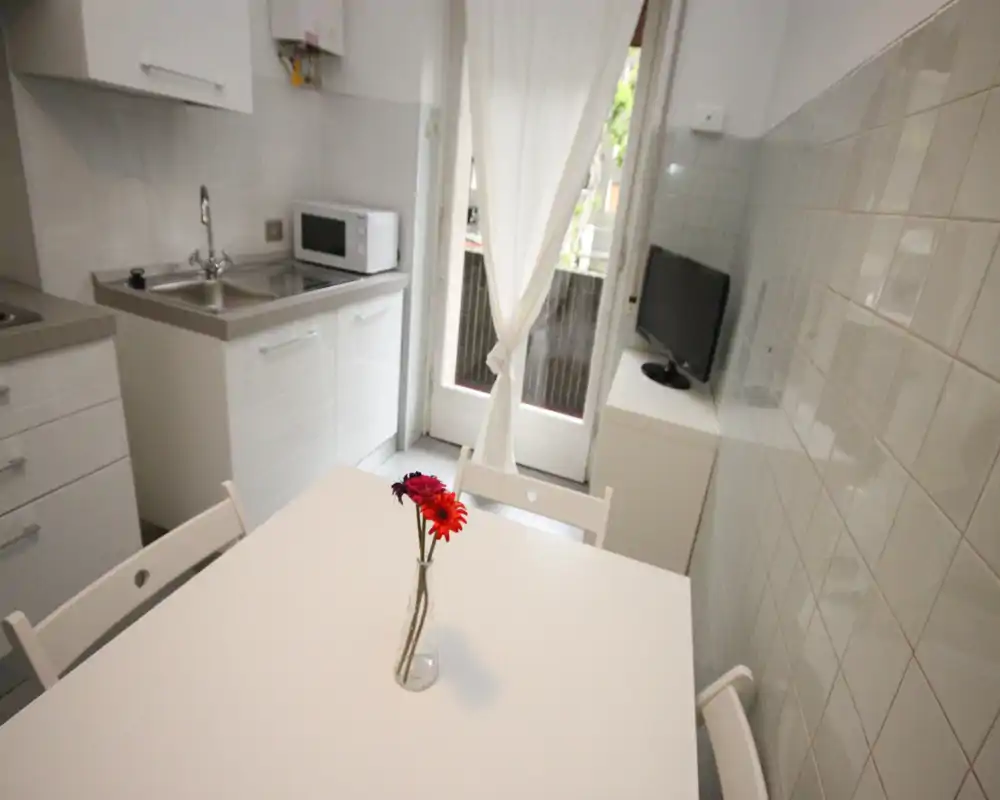 Cucina compatta e luminosa con tavolo bianco, lavello e microonde; porta finestra che introduce luce naturale e piccoli fiori decorativi che rendono l'ambiente accogliente.