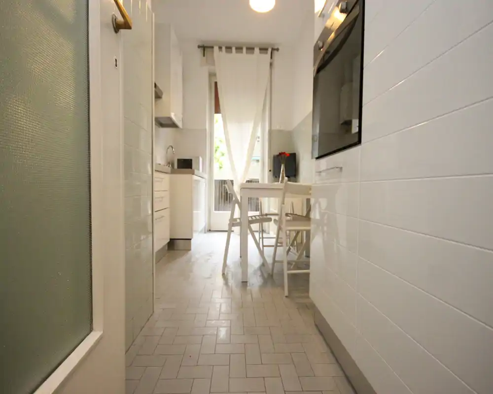 Cucina luminosa e stretta con un piccolo tavolo da pranzo e sedie, mobili bianchi e porta a vetro che lascia entrare luce naturale — ambiente ordinato e arioso, ideale per coppie o single.