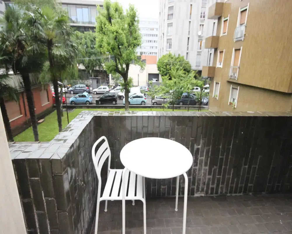 Piccolo balcone con tavolino e sedia bianchi, affacciato su un cortile verde e la strada — ideale per un caffè mattutino.