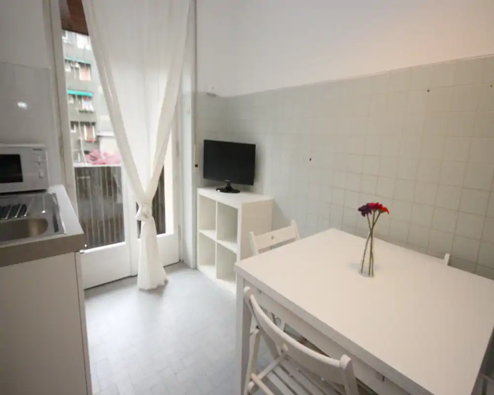 Cucina compatta e luminosa con piccolo tavolo da pranzo, mobili bianchi e porta finestra sul balcone; semplice, ordinata e ben illuminata per i pasti quotidiani.