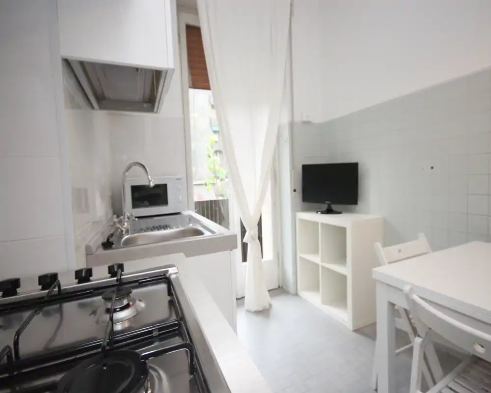 Cucina compatta e luminosa con piano cottura a gas in primo piano, lavello in acciaio e microonde, piccolo tavolo da pranzo e TV su scaffale; tonalità bianche e tenda leggera per luce naturale.