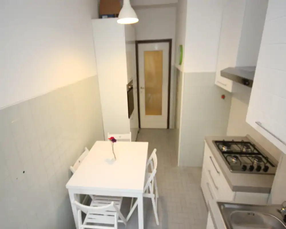 Cucina compatta e ben organizzata con mobili bianchi, piano cottura a gas a quattro fuochi, lavello e un piccolo tavolo con sedie — luminosa e ordinata, ideale per un appartamento in città.