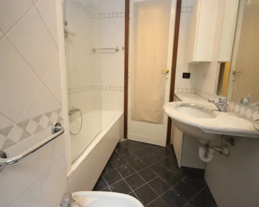 Bagno compatto con vasca/doccia, lavabo con piano in marmo e specchio a parete. Piastrelle in tinta neutra e illuminazione naturale moderata.