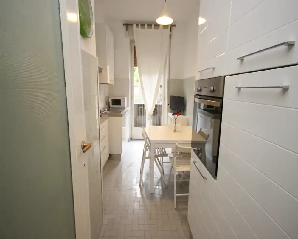 Cucina moderna e luminosa con forno incassato, mobili bianchi e un piccolo angolo pranzo vicino alla finestra — ordinata e ben illuminata, ideale per affitti brevi.