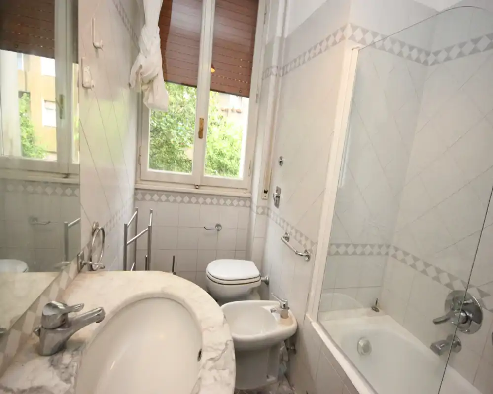 Bagno compatto e luminoso con piano lavabo in marmo, bidet, WC e vasca con parete in vetro sotto una grande finestra.