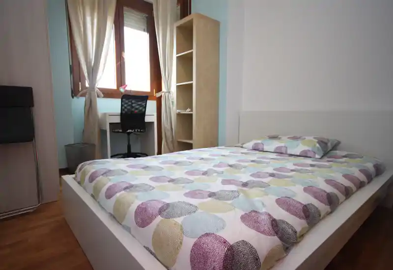 Dormitorio moderno y luminoso con cama doble en primer plano, edredón estampado y un pequeño escritorio junto a la ventana que aporta luz natural.