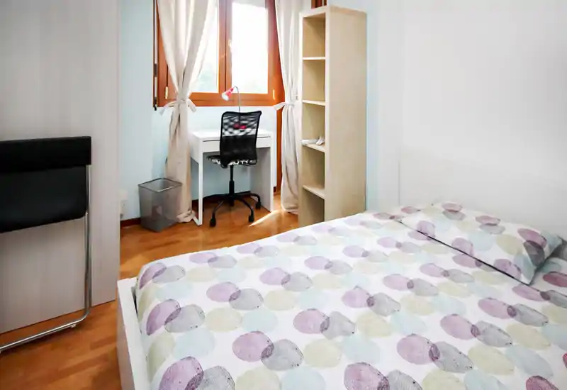 Dormitorio luminoso y minimalista con cama doble estampada, escritorio junto a la ventana y estantería abierta; líneas sencillas y luz natural que aportan sensación de amplitud.