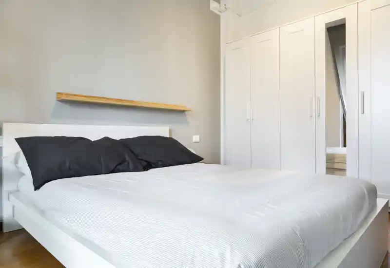 Camera da letto luminosa e minimal con un ampio letto bianco, cuscini neri e un armadio a muro bianco. L'ambiente appare ordinato e arioso con toni neutri e molta luce naturale.