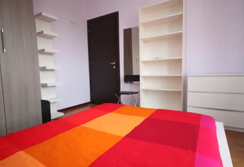 Camera da letto luminosa e ordinata con un grande letto colorato in primo piano, mensole semplici e un comò lungo le pareti — spazio notte moderno e minimale ideale per un annuncio.