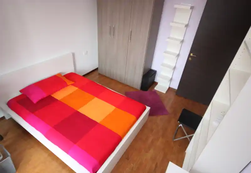 Camera da letto luminosa e ben arredata con un letto matrimoniale colorato, pavimento in legno e armadio a muro; dettagli accoglienti come un piccolo tappeto e mensole.