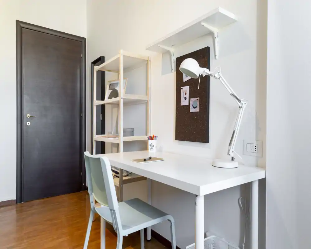 Bright, minimal workspace with a white desk, adjustable desk lamp, shelving unit and a simple chair — ideal for a focused home office or study corner.