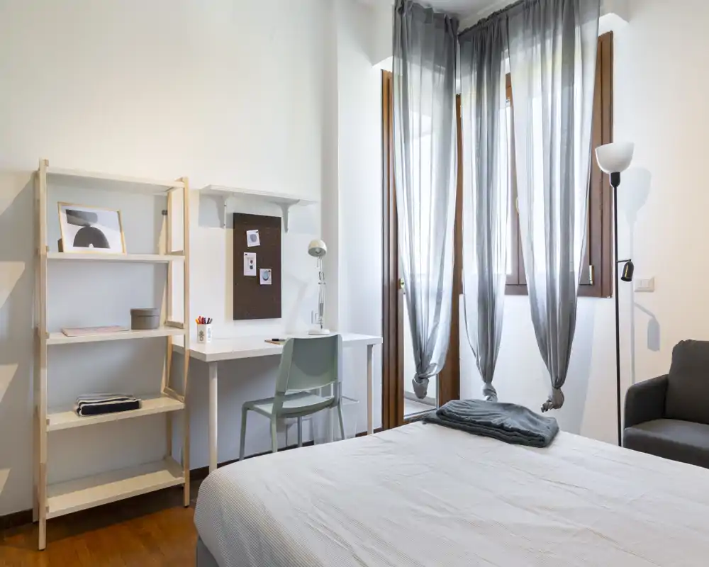 Bright, modern bedroom with a neatly made bed, a small desk and shelving, and large windows with sheer curtains letting in natural light.
