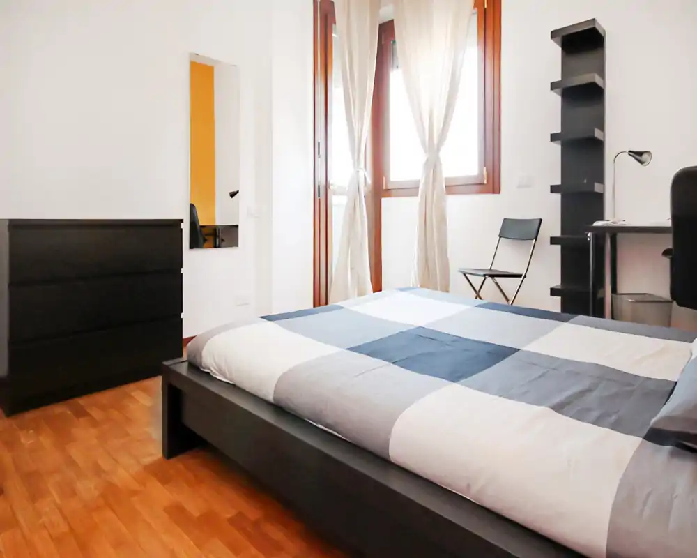 Bright modern bedroom with a low platform bed dressed in a gray-and-white check duvet, wooden floors and natural light from a large window with sheer curtains; includes dresser, desk and shelving for a minimalist, functional stay.