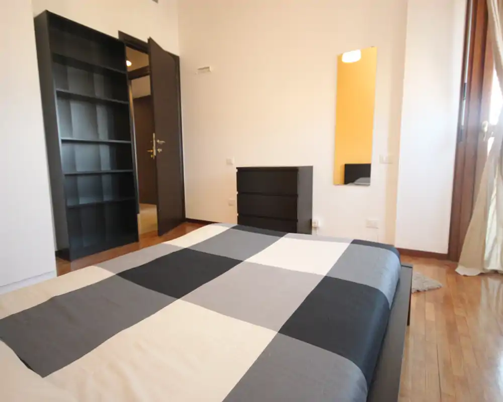 Bright, modern bedroom with a large double bed covered by a geometric duvet, wooden floors and minimal black furniture including a dresser and shelving unit. Natural light enters from a window/door on the right, creating a clean and airy atmosphere suitable for rental listings.