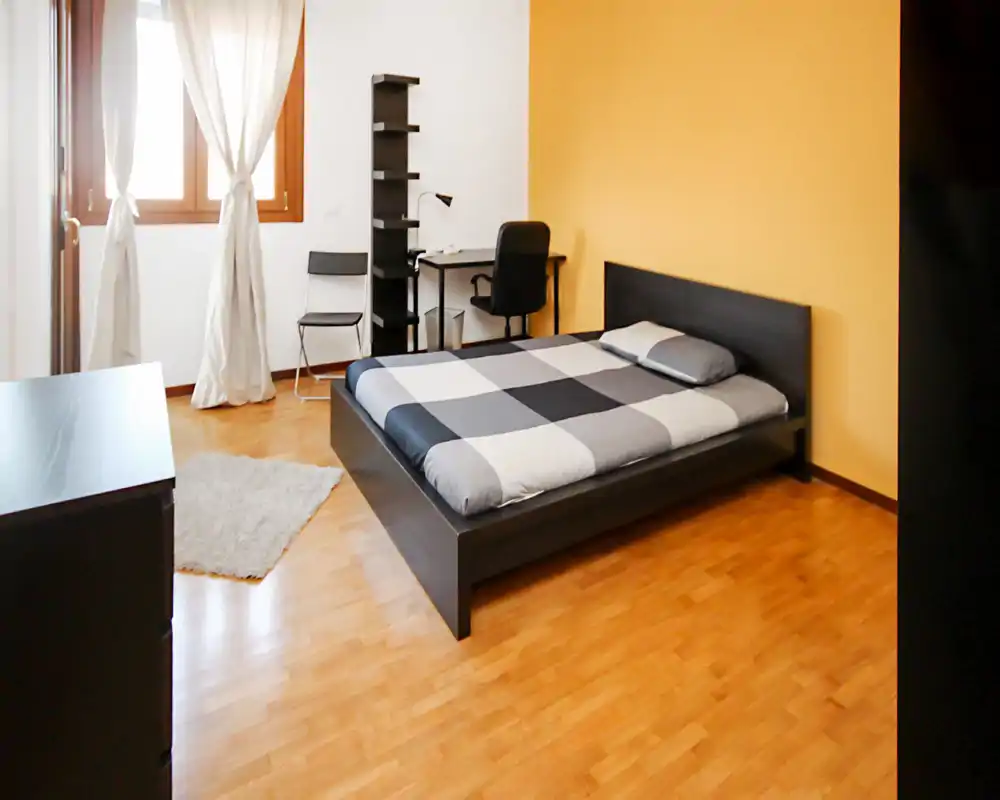 Bright, modern bedroom with a low-profile double bed, patterned grey bedding and a warm yellow accent wall. The room includes a small desk and shelving, wood floors and a window with light curtains providing natural light.