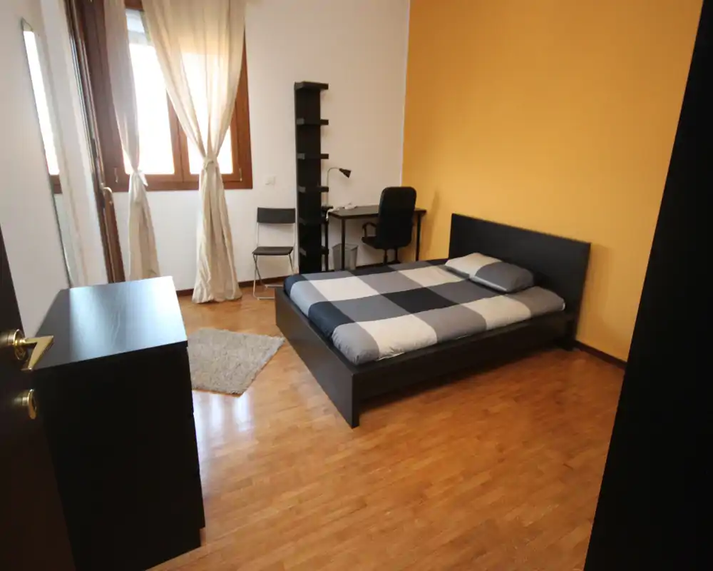 Bright, neatly staged bedroom with a full bed, wood flooring, a desk by the window and a warm yellow accent wall — ideal for single occupancy.