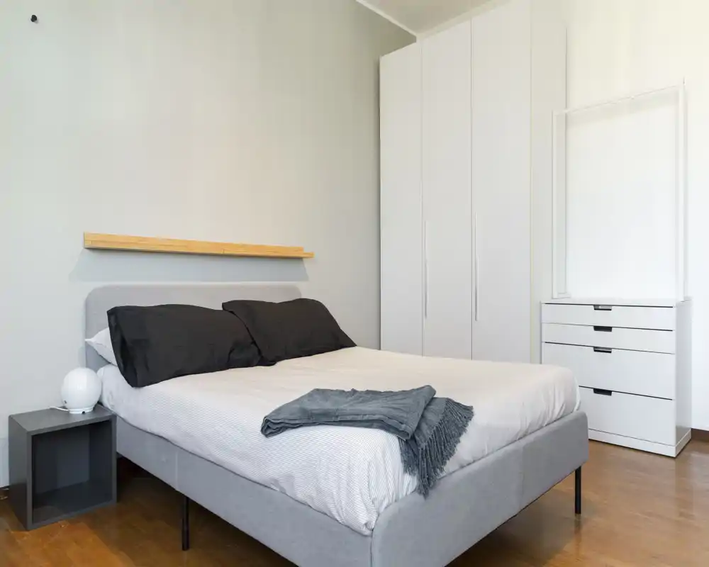 Minimalist, bright bedroom with a grey upholstered double bed, dark pillows and a folded throw on top; white wardrobe and chest of drawers on the right provide ample storage.