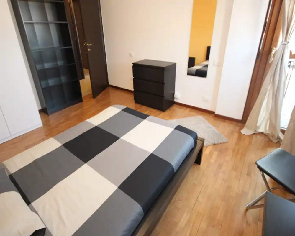 Bright modern bedroom with a large double bed covered in a geometric duvet, wooden floors, and simple black furniture. Natural light enters from the window creating a warm, welcoming atmosphere.