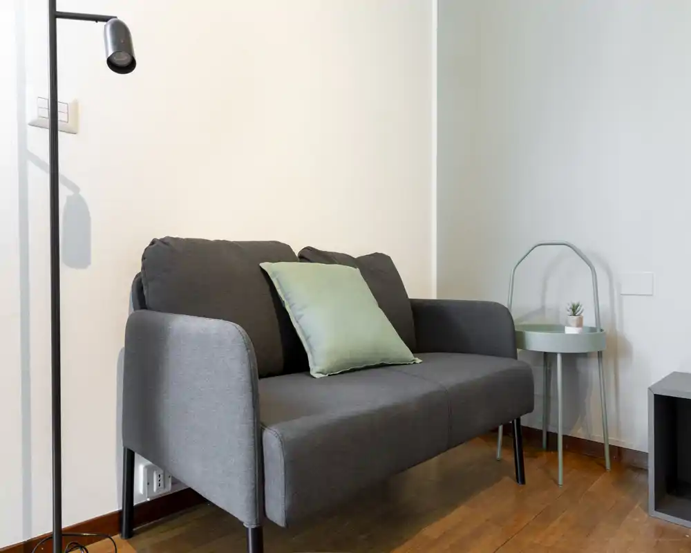 Compact living area with a two-seater grey sofa, green accent cushion and a small round side table — bright, minimal and comfortable for relaxing.