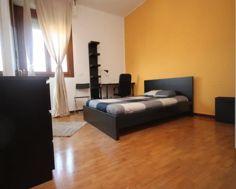 Tidy single bedroom with a low dark wood bed, warm yellow accent wall and natural light from a window. The room features a desk and shelving, polished wooden floors and simple, modern furnishings.