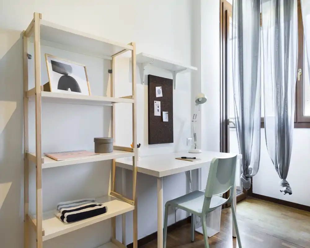 Bright, minimal study corner with a white desk, shelving unit and a light chair by a window letting in natural light — ideal for working or studying.