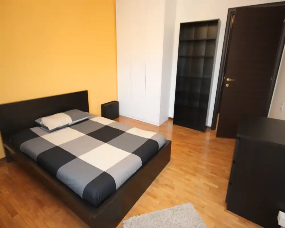 Bright, modern bedroom with a large low-profile bed, wardrobe and shelving. Warm accent wall and wooden floors create a cozy, well-lit space ideal for rentals.