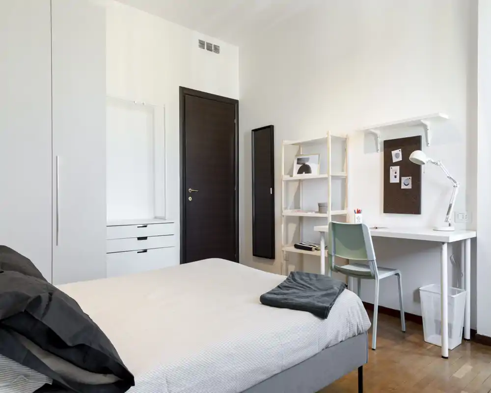 Bright, minimal bedroom with a neatly made double bed, a small desk and shelving unit — ideal for a student or professional seeking a tidy workspace in the room.