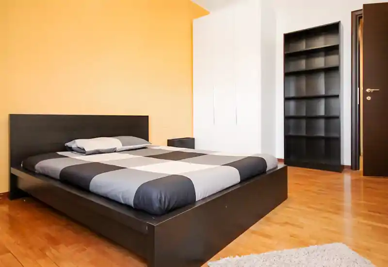 Spacious, modern bedroom with a low-profile dark wood bed and contrasting warm accent wall. Clean parquet flooring, a white wardrobe and open shelving complete the minimal, bright room.