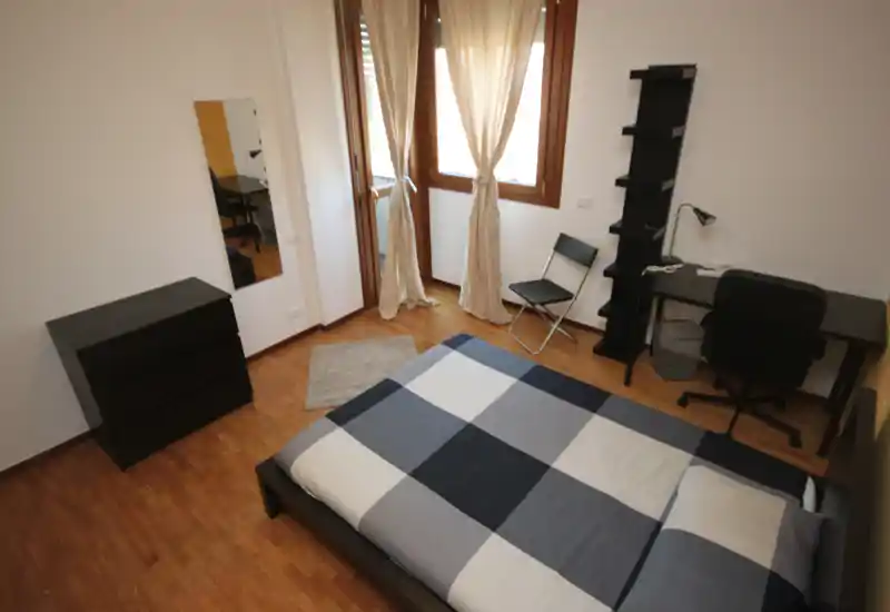 Bright, simply styled bedroom with a double bed centered in the frame, wooden floors, a desk and shelving by the window and soft curtains providing natural light.