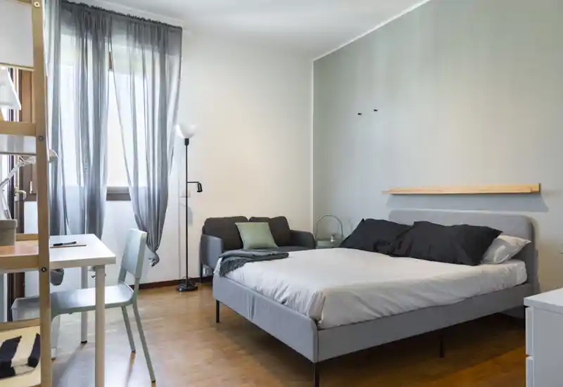 Bright, modern bedroom with a double bed, small sofa and desk area; soft grey tones and natural light make it welcoming and functional for stays or long-term rentals.