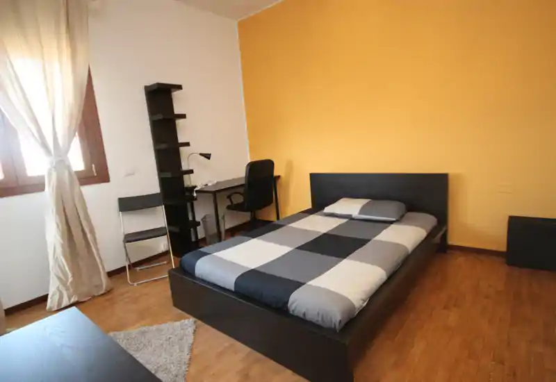 Bright, modern single bedroom with a double bed in a dark wood frame, desk and shelving by the window and warm yellow accent wall.