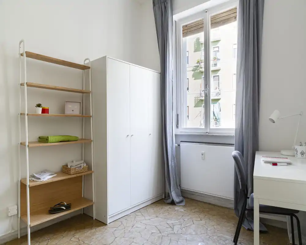 Minimal, bright bedroom corner with wardrobe, shelving, desk and a large window letting in natural light; simple, functional and well-lit space ideal for a single occupant.