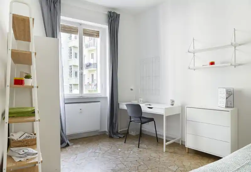 Bright, minimal bedroom with a small visible bed corner, a desk by the window and white modern storage—ideal for a single tenant seeking a clean, well-lit space.