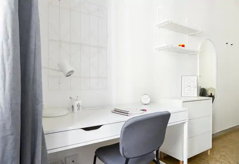 Bright, minimal workspace with a white desk, grey chair, small chest of drawers and simple wall shelving — ideal for a compact home office corner.