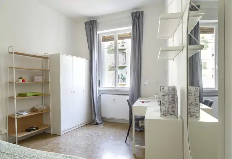 Bright, simply furnished bedroom with a bed at the corner, wardrobe, shelving and a small desk by the window letting in natural light.