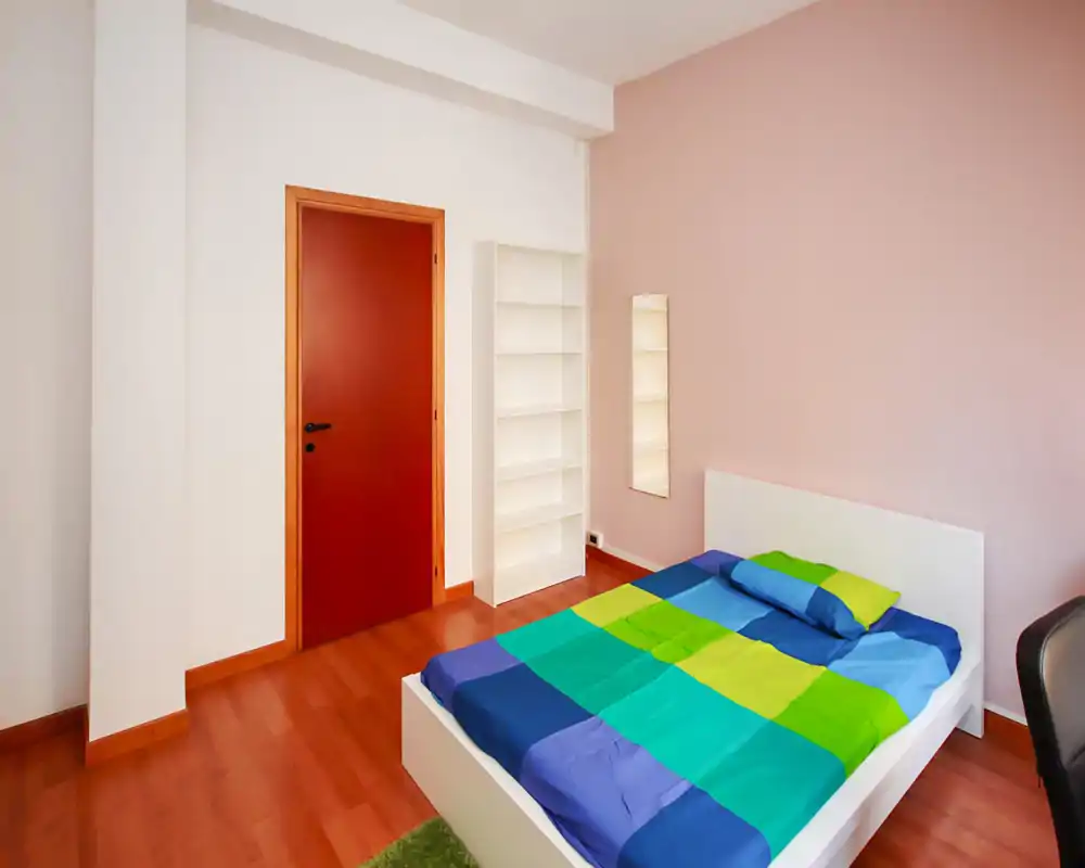 Bright, simply styled bedroom with a white low bed and colorful bedding, soft pink accent wall, and a slim shelving unit beside a full-length mirror.