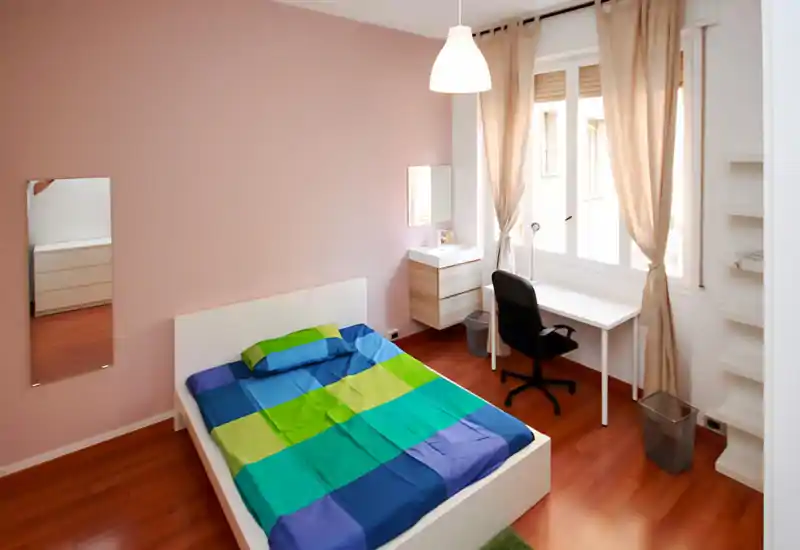 Bright, modern bedroom with a low white bedframe, colorful blue-green bedding, a desk by a large window and warm wooden floors — ideal for students or professionals.