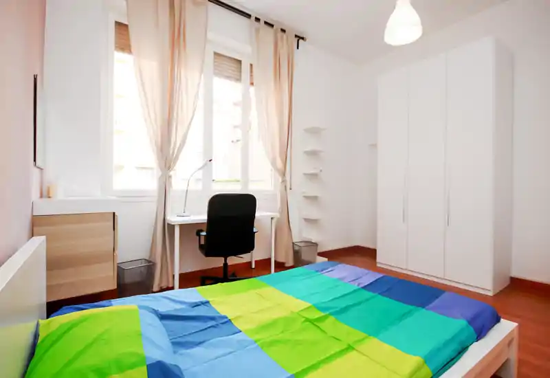 Bright, modern bedroom with a colorful double bed in the foreground, a window-side desk and chair, and a large white wardrobe. Natural light and minimalist decor create an airy, welcoming space ideal for rest and remote work.