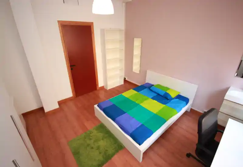 Bright, neatly arranged bedroom with a double bed dressed in a colorful duvet, a small green rug and simple shelving; well-lit and ready for guests.