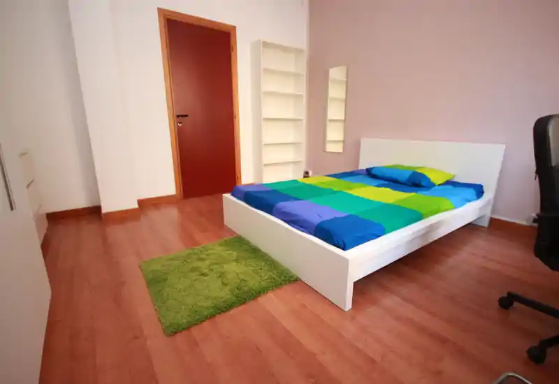 Bright, modern bedroom with a white platform bed, colorful duvet and a small green rug; clean wooden floor and minimal shelving create a fresh, uncluttered look.