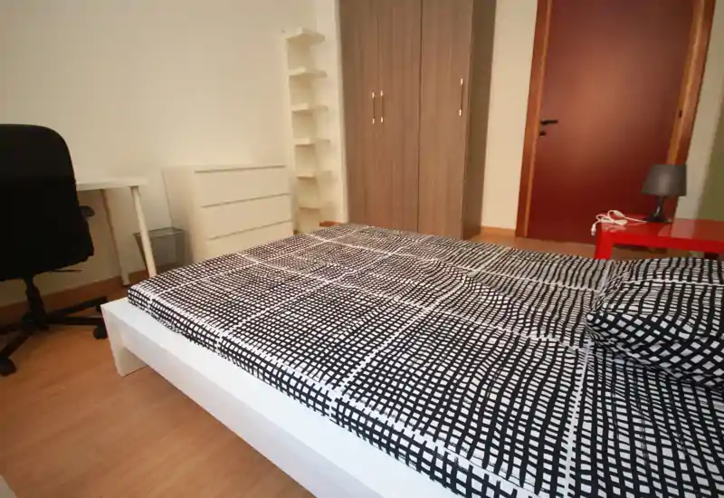 Dormitorio moderno con cama baja blanca con ropa de cama en blanco y negro, armario y escritorio al fondo; espacio luminoso, limpio y de mobiliario sencillo, ideal para alquileres.