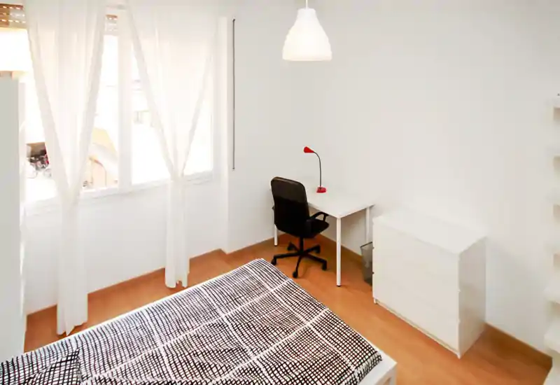 Dormitorio luminoso y minimalista con gran ventana, ropa de cama con estampado y pequeño escritorio con lámpara — ideal para una estancia compacta y funcional.