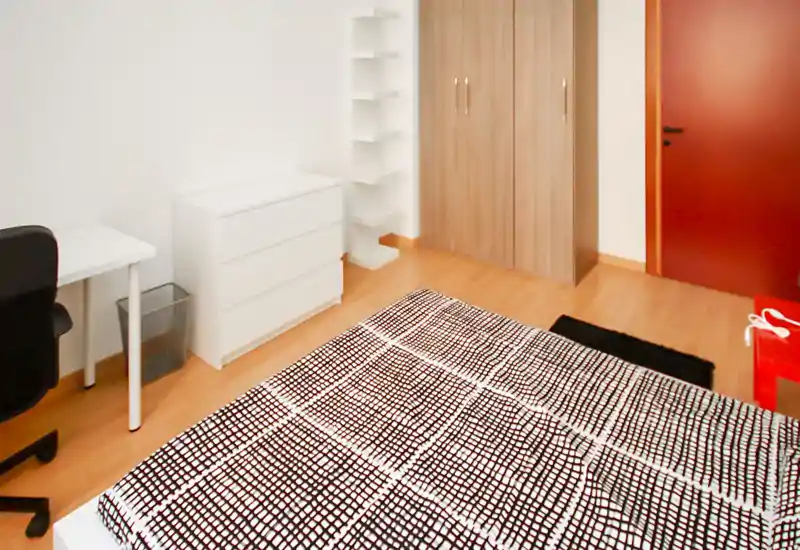 Dormitorio luminoso y con mobiliario sencillo: colcha estampada, cómoda blanca, armario y pequeño escritorio — ideal para una persona.