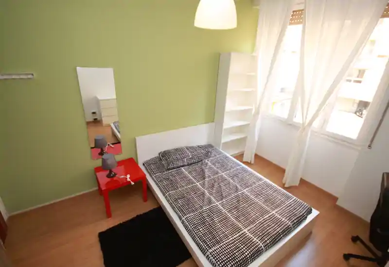 Dormitorio individual moderno y luminoso con estructura de cama blanca, ropa de cama estampada, mesita de noche roja con lámpara, estantería y grandes ventanas que aportan mucha luz natural.