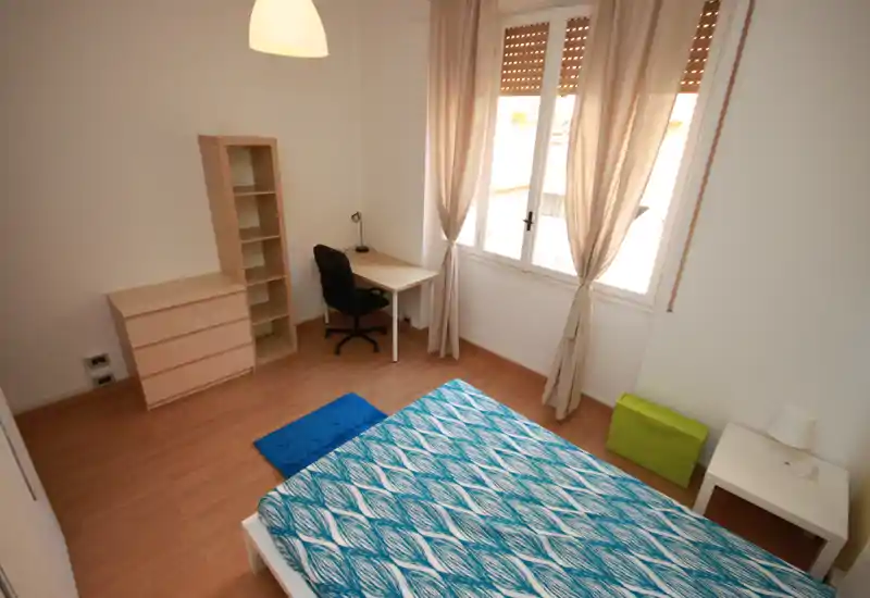 Bright, minimally furnished bedroom with a double bed, desk and shelving next to a large window letting in abundant natural light.